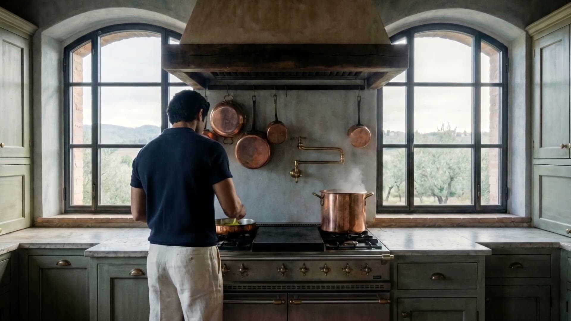 A cook at a range in a sunlit Mediterranean kitchen, copper pans on the wall between two arched windows