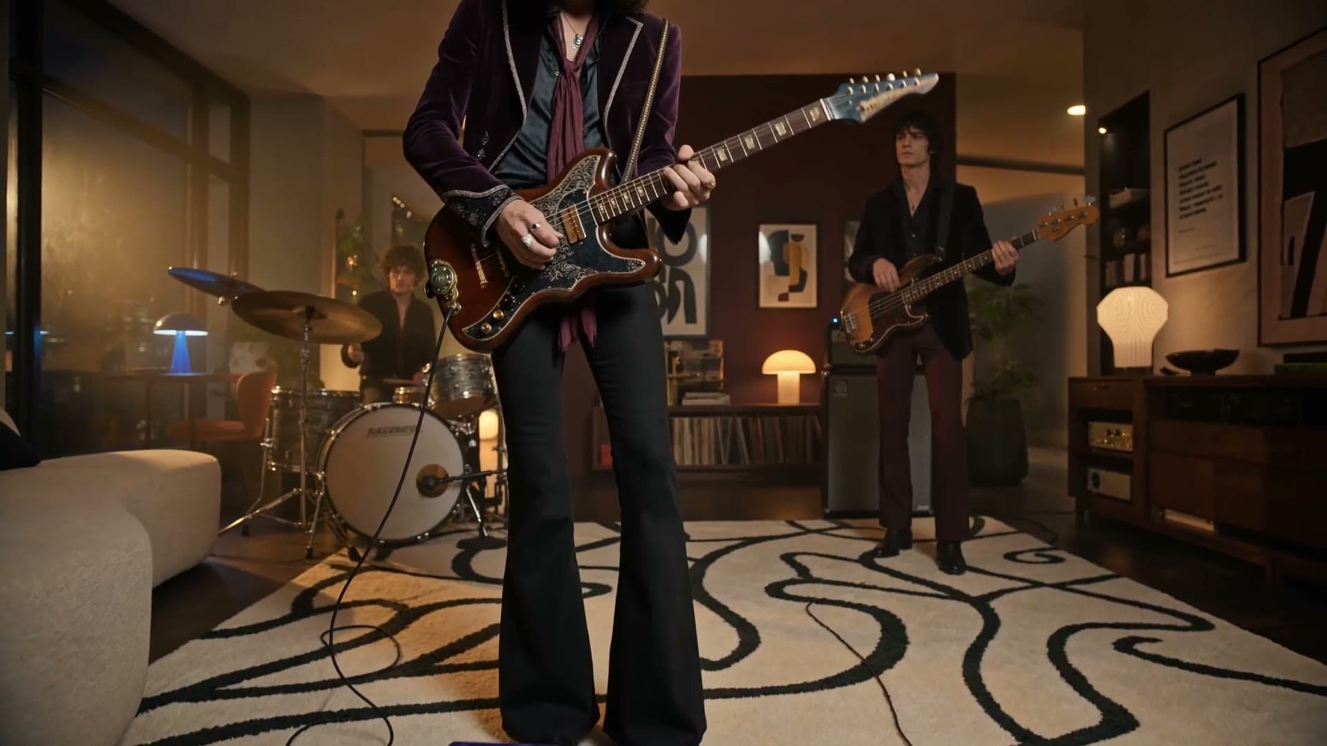 A guitarist mid-performance in a warmly lit living room, drummer and bassist behind